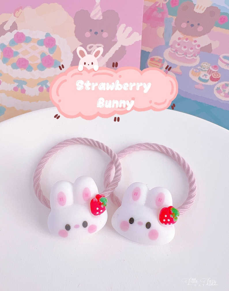 May include: Two white bunny hair ties with pink cheeks and a red strawberry accent. The hair ties have a pink rope-like band. The image includes a pink cloud-shaped sign that says "Strawberry Bunny".