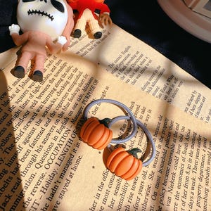 Pode incluir: Duas presilhas de cabelo em forma de ab&oacute;bora laranja e estatuetas de Halloween em um fundo de papel vintage. As estatuetas incluem um esqueleto e uma lanterna. A imagem tem um tema festivo e sazonal.