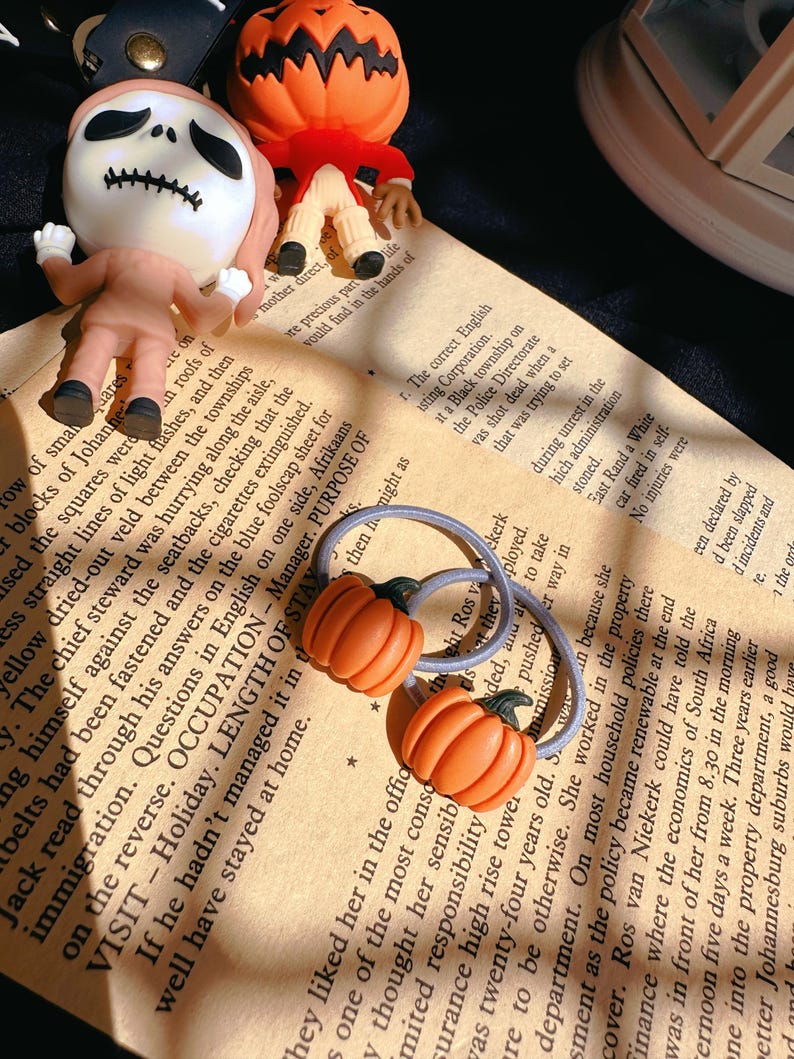 Pode incluir: Duas presilhas de cabelo em forma de ab&oacute;bora laranja e estatuetas de Halloween em um fundo de papel vintage. As estatuetas incluem um esqueleto e uma lanterna. A imagem tem um tema festivo e sazonal.