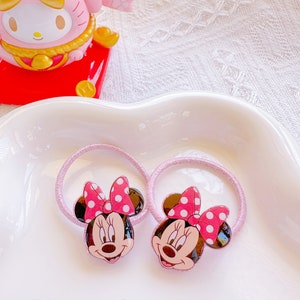 May include: Two pink hair ties featuring Minnie Mouse faces with red polka dot bows. The text "Minnie 1" is displayed in pink and purple. The hair ties are on a white surface.