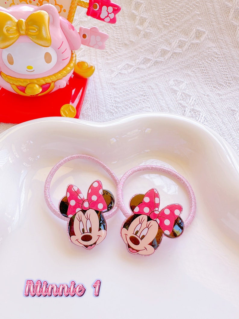 May include: Two pink hair ties featuring Minnie Mouse faces with red polka dot bows. The text "Minnie 1" is displayed in pink and purple. The hair ties are on a white surface.