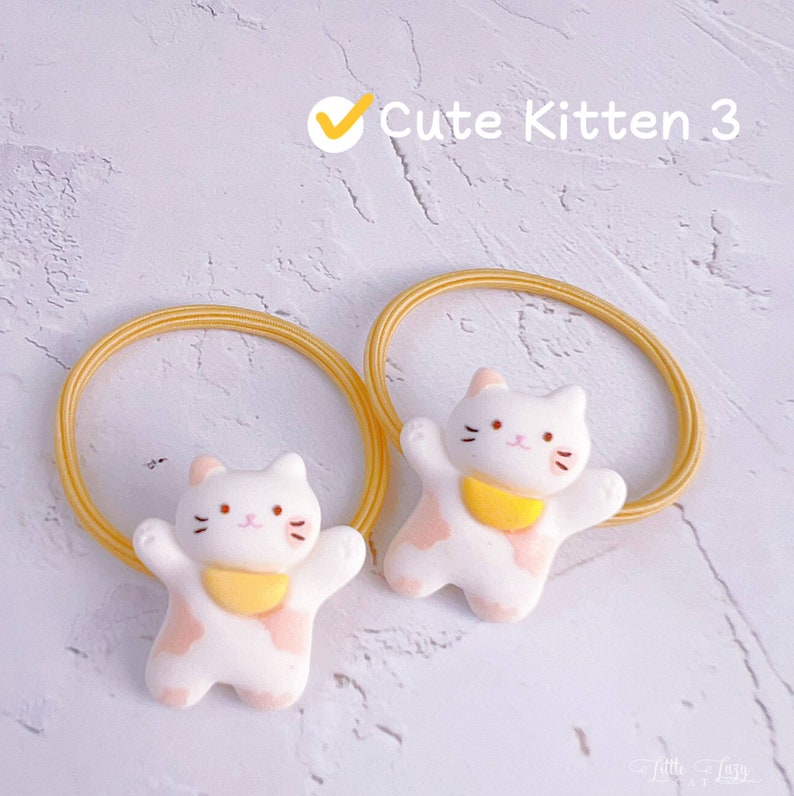Puede incluir: Dos coleteros con abalorios de gato blanco con detalles rosas y baberos amarillos. Las gomas para el pelo son amarillas. El texto "Cute Kitten 3" es visible.