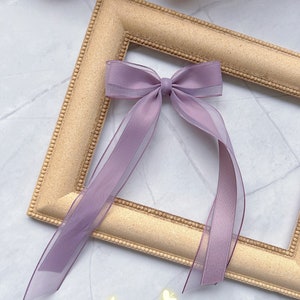 May include: A lilac bow with long, sheer ribbons, displayed on a gold-coloured picture frame. The text "Style 1" is visible at the bottom. The bow is a delicate hair accessory.