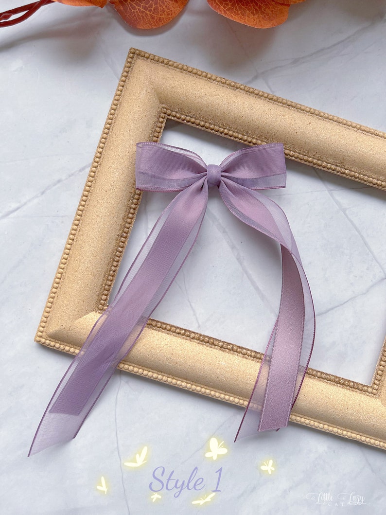 May include: A lilac bow with long, sheer ribbons, displayed on a gold-coloured picture frame. The text "Style 1" is visible at the bottom. The bow is a delicate hair accessory.