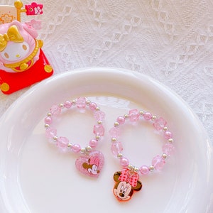 May include: Two pink beaded bracelets with silver accents and Minnie Mouse charms. One charm is a heart with Minnie's image, the other is a Minnie Mouse head. The bracelets are on a white plate with a pink and yellow figurine.