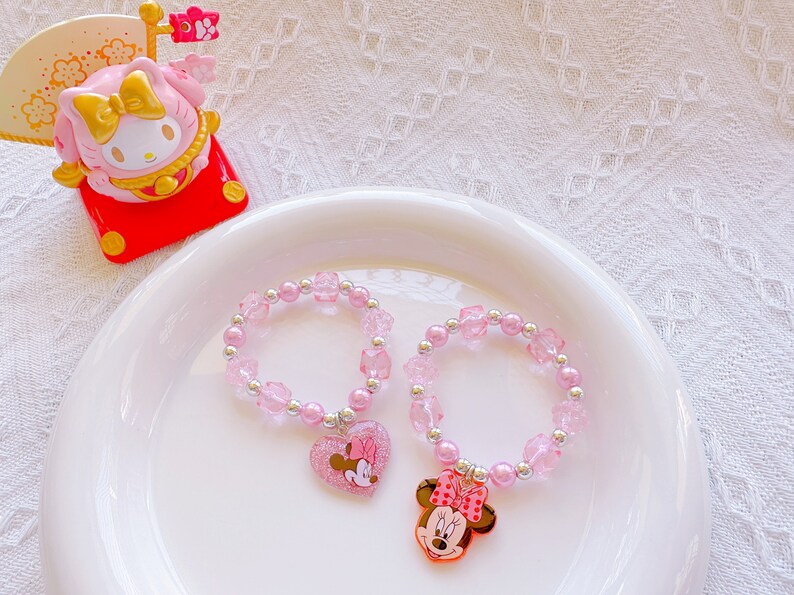 May include: Two pink beaded bracelets with silver accents and Minnie Mouse charms. One charm is a heart with Minnie's image, the other is a Minnie Mouse head. The bracelets are on a white plate with a pink and yellow figurine.