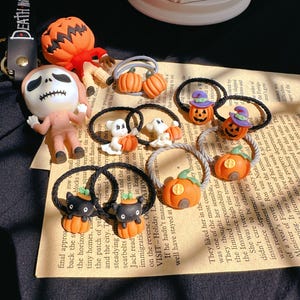 Pode incluir: Uma cole&ccedil;&atilde;o de el&aacute;sticos de cabelo e figuras com tema de Halloween. Os el&aacute;sticos de cabelo apresentam desenhos de ab&oacute;bora, fantasma e gato preto. As figuras incluem um espantalho com cabe&ccedil;a de ab&oacute;bora e uma figura de esqueleto. O texto "DEATH ME" &eacute; vis&iacute;vel.