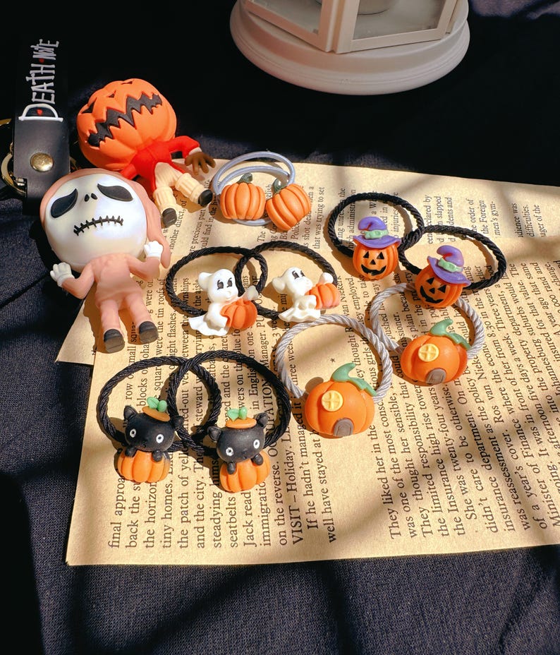 Pode incluir: Uma cole&ccedil;&atilde;o de el&aacute;sticos de cabelo e figuras com tema de Halloween. Os el&aacute;sticos de cabelo apresentam desenhos de ab&oacute;bora, fantasma e gato preto. As figuras incluem um espantalho com cabe&ccedil;a de ab&oacute;bora e uma figura de esqueleto. O texto "DEATH ME" &eacute; vis&iacute;vel.