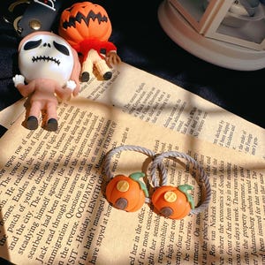 Pode incluir: Acess&oacute;rios com tema de Halloween, incluindo um chaveiro Jack Skellington, uma figura com cabe&ccedil;a de ab&oacute;bora e dois el&aacute;sticos de cabelo de ab&oacute;bora laranja. Os itens est&atilde;o em um fundo de papel vintage, evocando uma atmosfera festiva.