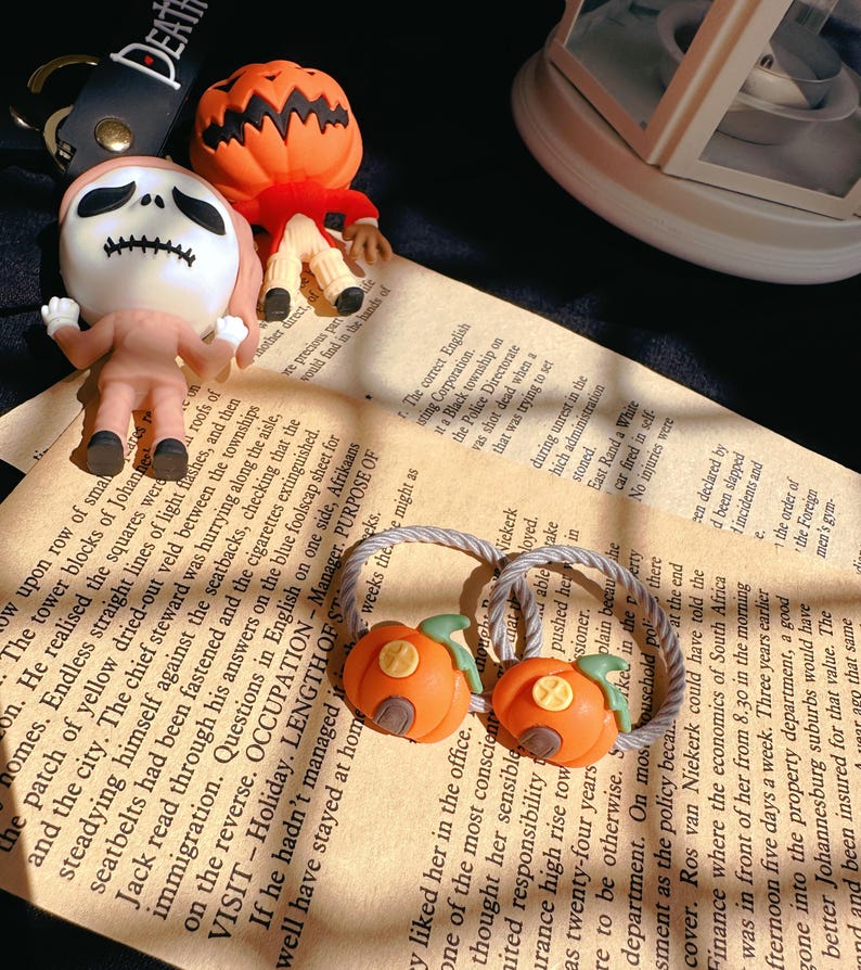 Pode incluir: Acess&oacute;rios com tema de Halloween, incluindo um chaveiro Jack Skellington, uma figura com cabe&ccedil;a de ab&oacute;bora e dois el&aacute;sticos de cabelo de ab&oacute;bora laranja. Os itens est&atilde;o em um fundo de papel vintage, evocando uma atmosfera festiva.