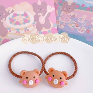 May include: Two brown bear-shaped hair ties with pink cheeks and white noses sit on a white plate. The hair ties have brown elastic bands. The background includes a pink and purple illustration with the text "Cute Guinea Pig".