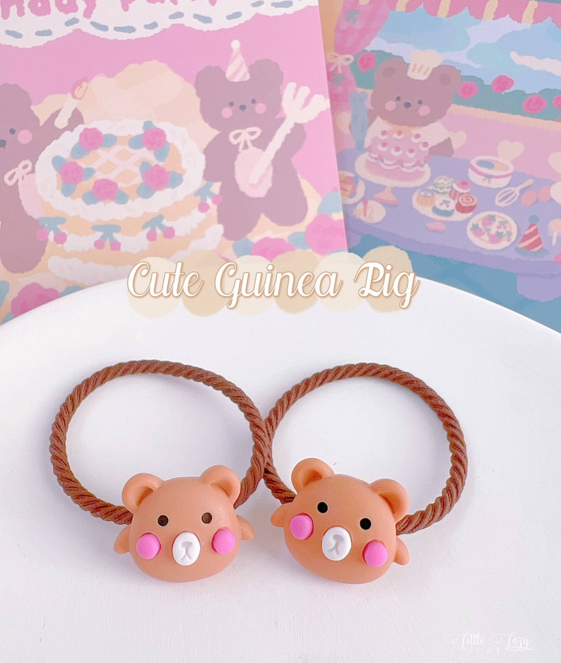 May include: Two brown bear-shaped hair ties with pink cheeks and white noses sit on a white plate. The hair ties have brown elastic bands. The background includes a pink and purple illustration with the text "Cute Guinea Pig".