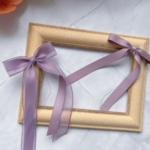 May include: A gold-coloured rectangular picture frame with a beaded edge. Two lavender bows with long, sheer ribbons are attached to the frame. The ribbons hang down, adding a delicate touch to the frame's design.