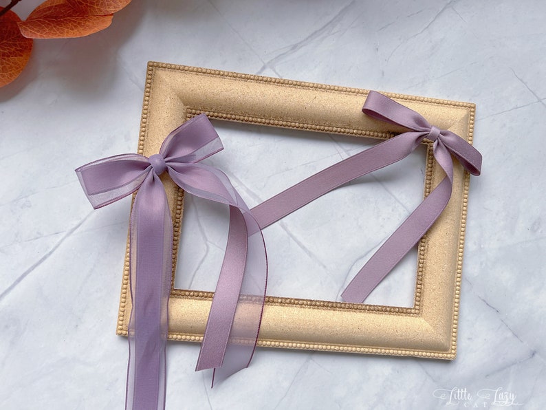 May include: A gold-coloured rectangular picture frame with a beaded edge. Two lavender bows with long, sheer ribbons are attached to the frame. The ribbons hang down, adding a delicate touch to the frame's design.