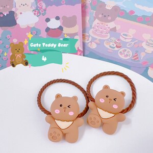May include: Two brown teddy bear hair ties with a braided brown elastic band. The bears have pink cheeks and a white bib. The image also includes a sign that says "Cute Teddy Bear".