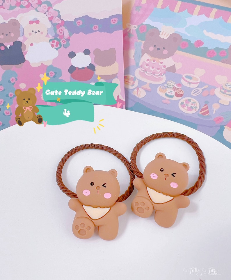 May include: Two brown teddy bear hair ties with a braided brown elastic band. The bears have pink cheeks and a white bib. The image also includes a sign that says "Cute Teddy Bear".