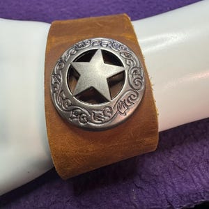 May include: A brown leather bracelet with a silver-colored star-shaped emblem. The emblem is circular with an embossed design around the star. The bracelet is shown on a white surface against a purple background.