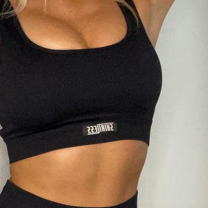 Op de afbeelding: Zwarte sportbeha met een ronde hals en een geribbelde textuur. De beha heeft een klein wit logo met de tekst "223TRINE" op de voorkant.