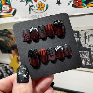 May include: A black card with six different nail designs. The designs include a black and white rose, a red and white rose, a black and white fang, a red and white fang, a black and white skull, and a red and white skull.