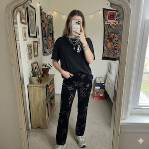 May include: A plain black short-sleeved t-shirt paired with black corduroy pants featuring an embroidered floral pattern in shades of red, pink, and green. The outfit is accessorized with layered silver necklaces and light-colored athletic sneakers.
