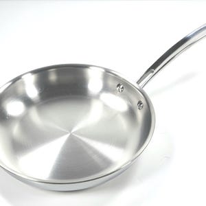 Puede incluir: Una sartén de acero inoxidable brillante con un mango largo y curvo. La sartén tiene una superficie reflectante y es redonda con un borde ligeramente inclinado. El mango está remachado y tiene un diámetro de aproximadamente 25 cm.