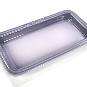 Puede incluir: Una bandeja rectangular de gres Le Creuset con un acabado brillante en color morado apagado. El plato presenta una base lisa y plana con bordes redondeados ligeramente elevados y un reborde. El nombre de la marca Le Creuset está grabado.