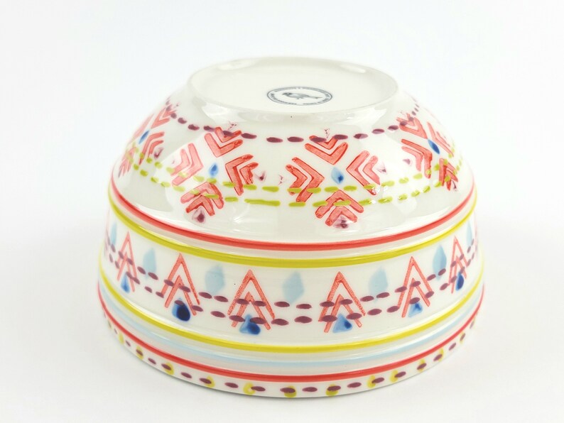 May include: White ceramic bowl with a colourful geometric pattern. The pattern features red, yellow, blue, and green colours. The bowl is perfect for serving snacks, salads, or desserts.