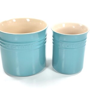 May include: Two Le Creuset ceramic utensil holders in a light blue color. The larger holder is on the left, and the smaller one is on the right. Both have a cream-colored interior and the Le Creuset logo.
