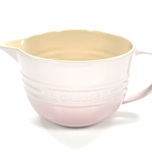 May include: A light pink Le Creuset mixing bowl with a handle and a pouring spout. The bowl has a cream-colored interior and the brand name is embossed on the side. The bowl is made of ceramic.