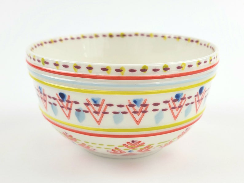 May include: A white ceramic bowl with a colourful geometric pattern. The pattern features red, yellow, blue, and purple accents. The bowl is ideal for serving snacks, salads, or desserts.