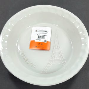May include: A light gray Le Creuset pie dish with a fluted edge. The dish features an embossed Eiffel Tower design and the Le Creuset logo. A product label is inside the dish.