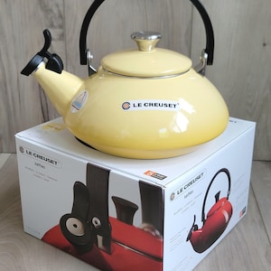 Może przedstawiać: Jasnożółty czajnik Le Creuset z czarnym uchwytem i wylewką, stojący na oryginalnym pudełku. Pudełko zawiera zdjęcia czajnika i logo Le Creuset. Czajnik ma srebrną pokrywę i czarny uchwyt.