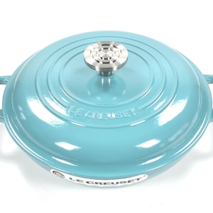 May include: A teal-colored Le Creuset Dutch oven with a matching lid. The pot has two loop handles and a silver-colored knob on top. The brand name is visible on the pot.
