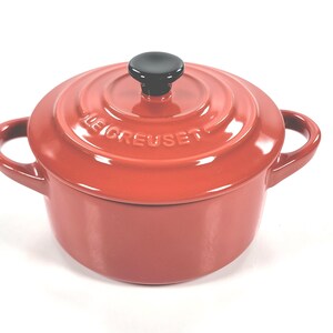 May include: A red enameled cast iron Le Creuset Dutch oven with a black knob on the lid. The pot has two loop handles and the brand name is embossed on the lid. The pot is on a white background.