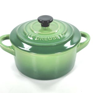 May include: A green Le Creuset casserole dish with a lid and two handles. The pot is a gradient of green, with a black knob on the lid. The brand name is embossed on the lid. This small pot is ideal for cooking and serving.