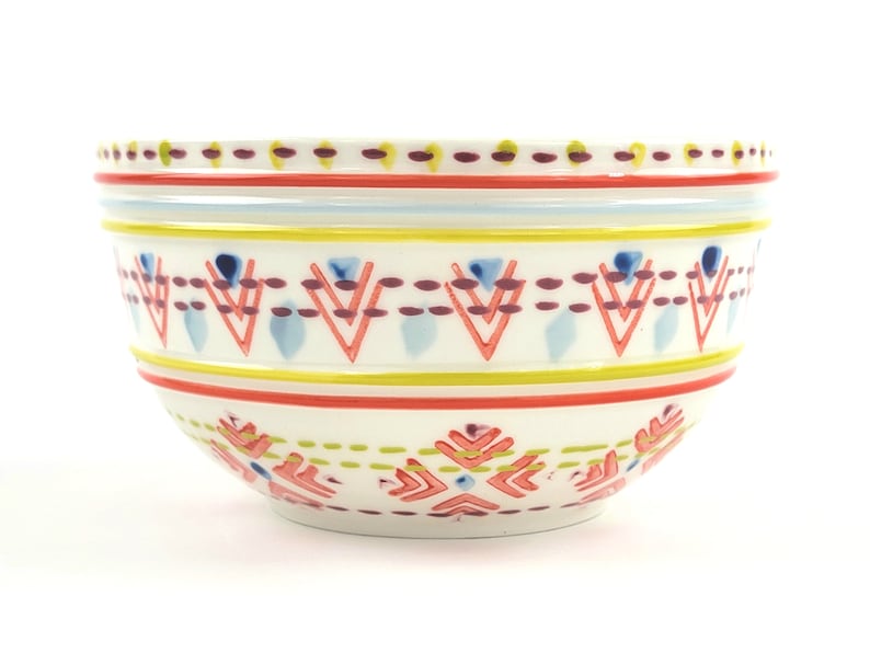 May include: A white ceramic bowl with a colourful geometric pattern. The pattern features red, yellow, blue, and purple accents. The bowl is ideal for serving snacks, salads, or desserts.