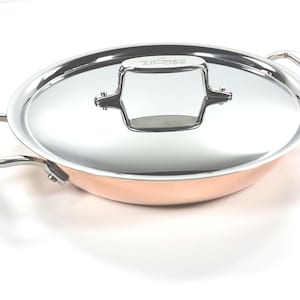 Puede incluir: Una sartén de color cobre con una tapa de acero inoxidable. La sartén tiene dos asas y un asa para la tapa. La tapa es de acero inoxidable y tiene un asa plateada. La sartén es redonda.