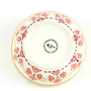 May include: A white ceramic bowl with a red, yellow and blue geometric pattern. The bowl is microwave and dishwasher safe and made in China. The bottom of the bowl has a small bird logo.