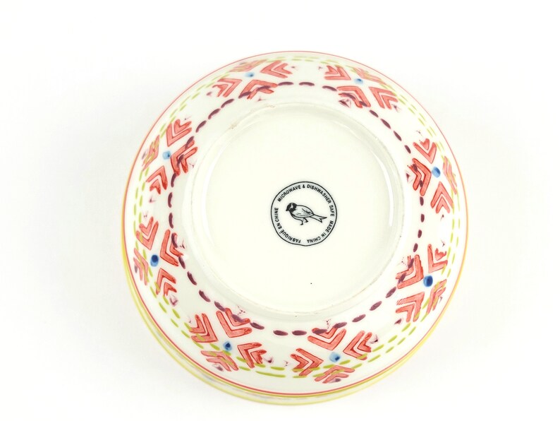 May include: A white ceramic bowl with a red, yellow and blue geometric pattern. The bowl is microwave and dishwasher safe and made in China. The bottom of the bowl has a small bird logo.