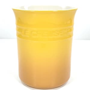 May include: A yellow Le Creuset utensil holder with a white interior. The cylindrical holder has a glossy finish and the brand name is embossed around the middle. The color gradient transitions from a bright yellow at the top to a darker yellow at the base.