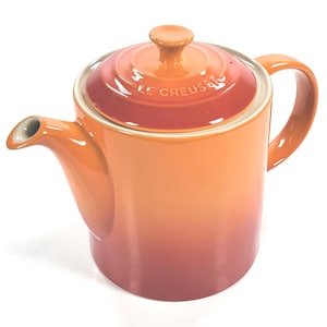 Pode incluir: Bule de cerâmica laranja e vermelho com tampa e alça. O bico é visível e a tampa tem a inscrição "LE CREUSET". O bule está sobre um fundo branco.