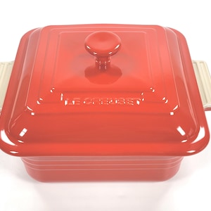 Puede incluir: Una olla cuadrada roja Le Creuset con tapa a juego y pomo redondo. La olla tiene asas color crema. El nombre de la marca está grabado en la tapa.