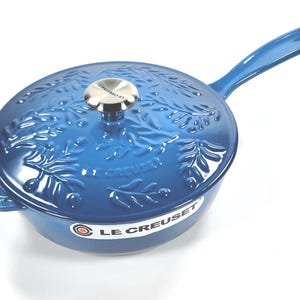 May include: A blue cast iron skillet with a lid. The skillet has a long handle and a lid with a silver knob. The lid and skillet are decorated with a leaf pattern. The brand name "Le Creuset" is visible on the side.