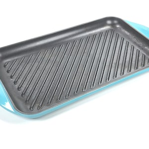 May include: A rectangular cast iron grill pan with a light blue enamel exterior and a dark grey interior. The pan has a ridged grilling surface and rounded handles. The brand name is visible on the handle.