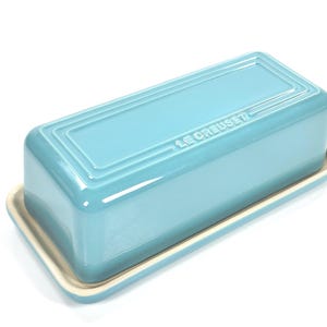May include: A light blue Le Creuset butter dish. The rectangular dish has a lid with a ridged design and sits on a matching base. The brand name is embossed on the lid. The dish is made of ceramic.