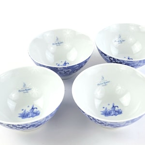 May include: Set of four white ceramic bowls with a blue and white floral pattern. The bowls feature the text "The Tale of Peter Rabbit" and a small image of Peter Rabbit.