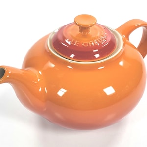 Puede incluir: Tetera de cerámica naranja con tapa a juego. La tapa presenta el nombre de la marca "Le Creuset" en letras blancas. La tetera tiene un asa curva y un pico para verter. Un artículo de menaje.