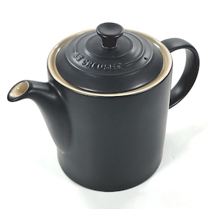 Puede incluir: Una tetera Le Creuset negra con tapa y asa a juego. La tapa tiene un borde dorado y un pomo negro. La tetera tiene un pico y está diseñada para preparar té. La tetera es de cerámica.