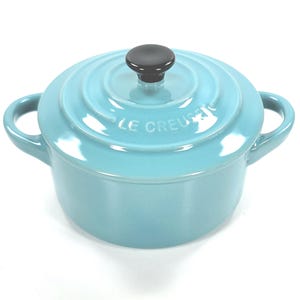 May include: A light blue Le Creuset Dutch oven with a matching lid and black knob. The small pot has two side handles and is made of enameled cast iron. The brand name is embossed on the lid.