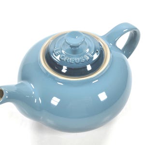 Może przedstawiać: Zakurzony niebieski dzbanek do herbaty z ceramiki z pasującą pokrywką z wytłoczonym napisem LE CREUSET. Dzbanek ma zaokrąglony korpus, zakrzywiony uchwyt i krótki dzióbek z beżową wewnętrzną krawędzią widoczną wokół pokrywki i wewnątrz dzióbka.